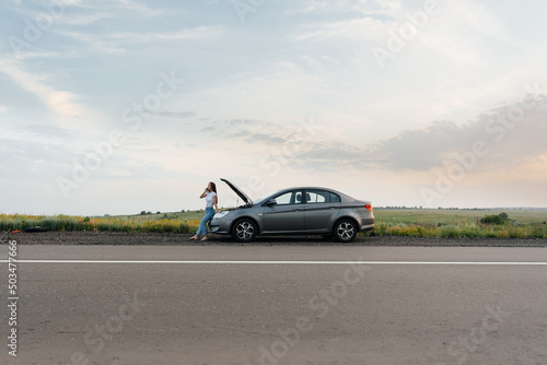 A young girl stands near a broken-down car in the middle of the highway during sunset and tries to call for help on the phone. Waiting for help. Car service. Car breakdown on road.