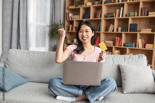 Overjoyed korean woman with laptop computer and credit card making YES gesture over big discount, sitting at home