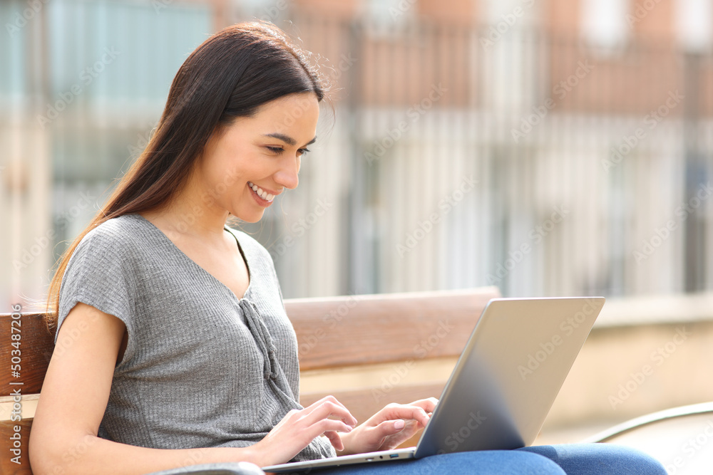 Happy woman using laptop sitting on bench in the street Happy woman using laptop sitting on bench in the street