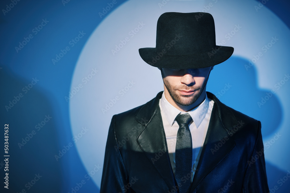 Portrait of handsome man in black coat hiding face behind his hat in the spotlight on studio background Portrait of handsome man in black coat hiding face behind his hat in the spotlight on studio background
