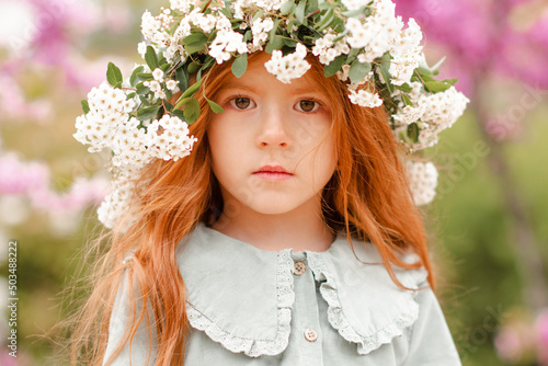 Cute little kid girl 3-4 year old with long curly red hair wear floral wreath and stylish rustic dress over nature background in garden outdoor. Springtime. Calm pretty baby toddler in meadow.