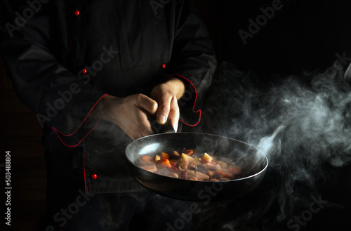Fototapeta Naklejka Na Ścianę i Meble -  The chef prepares food in a frying pan with steam on a black background. The concept of restaurant and hotel service. Asian cuisine