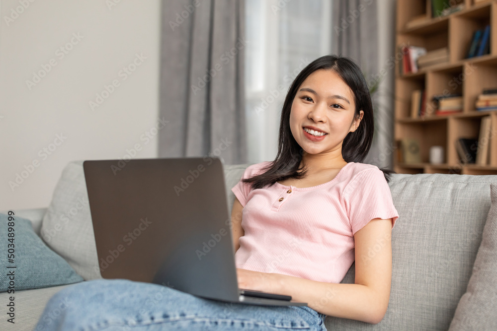 Portrait of happy asian lady sitting on sofa with laptop on laps and smiling at camera, having distance job Portrait of happy asian lady sitting on sofa with laptop on laps and smiling at camera, having distance job