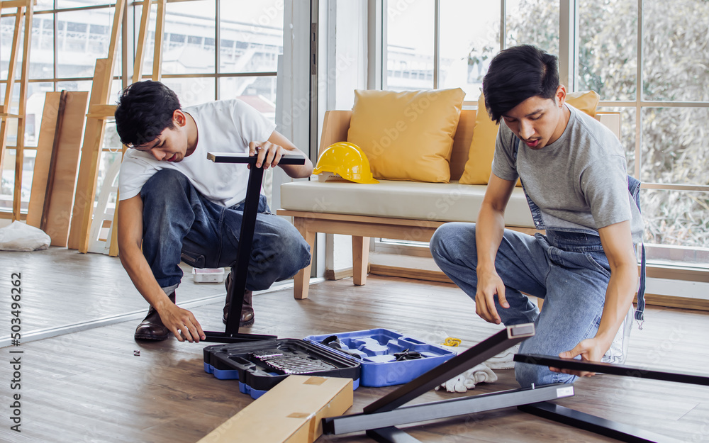 Two young Asian handsome men or friends helping each other, repairing or making DIY assembly furniture in living room at indoor home. Teamwork, Business Service, Interior and Industry Concept. Two young Asian handsome men or friends helping each other, repairing or making DIY assembly furniture in living room at indoor home. Teamwork, Business Service, Interior and Industry Concept.