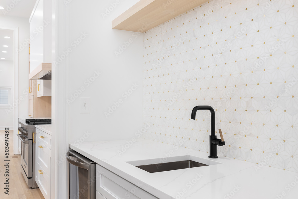 Sink detail shot with a black and gold faucet, white cabinet and marble countertop, and a white and gold mosaic tile looking out towards a modern kitchen. - obrazy, fototapety, plakaty 