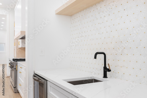 Sink detail shot with a black and gold faucet, white cabinet and marble countertop, and a white and gold mosaic tile looking out towards a modern kitchen.