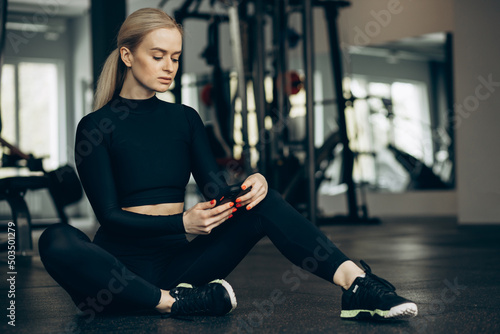 Obraz na plátně Woman sitting on the floor at the gym and using mobile phone