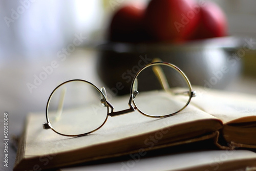 Silver bowl filled with red apples, open book and reading glasses on the table. Selective focus.
