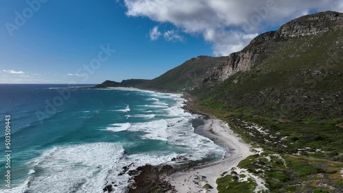Aerial dolly shot, of waves crashing on a beach below rocky cliffs on a bright sunny day with partly cloudy sky, Misty Cliffs near Cape Town South Africa,