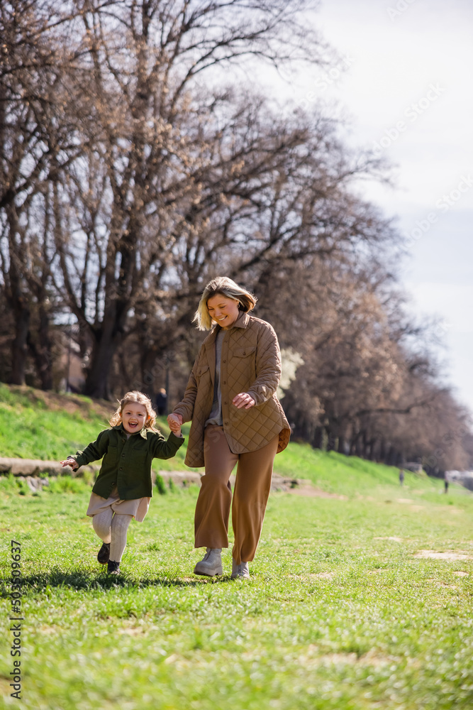 full length view of pleased mother and daughter in stylish clothes walking in green park. full length view of pleased mother and daughter in stylish clothes walking in green park.