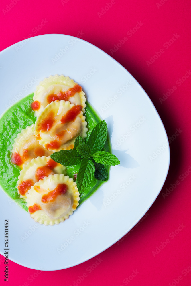 Italian pasta ravioli with burrata filling with tomato and pea cream on white plate shot from above with red background in vertical shot Italian pasta ravioli with burrata filling with tomato and pea cream on white plate shot from above with red background in vertical shot