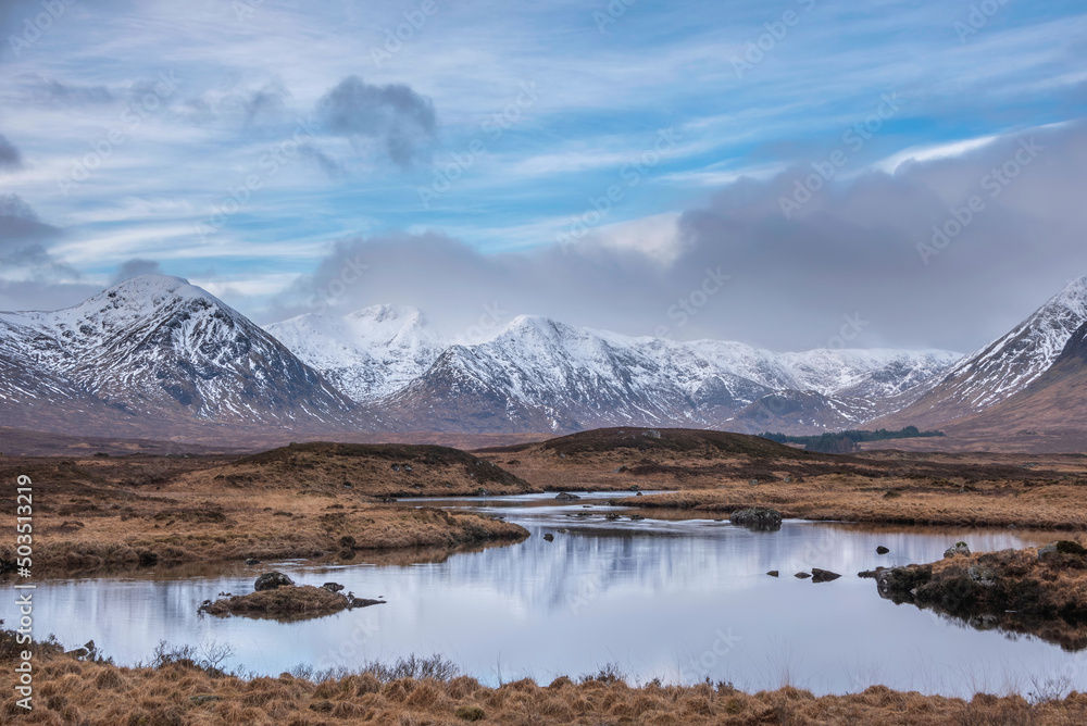 Beautiful Winter panorama landscape image of mountain range and peaks viewed from Loch Ba in Scottish Highlands with dramatic clouds overhead