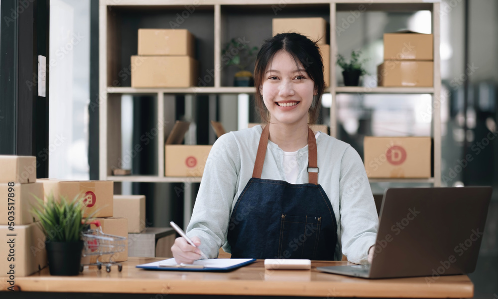 A portrait of a young Asian woman, e-commerce employee sitting in the office full of packages in the background write note of orders and a calculator, for SME business ecommerce and delivery business. A portrait of a young Asian woman, e-commerce employee sitting in the office full of packages in the background write note of orders and a calculator, for SME business ecommerce and delivery business.