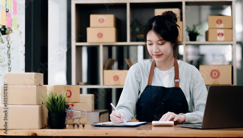 A portrait of a young Asian woman, e-commerce employee sitting in the office full of packages in the background write note of orders and a calculator, for SME business ecommerce and delivery business. © wichayada