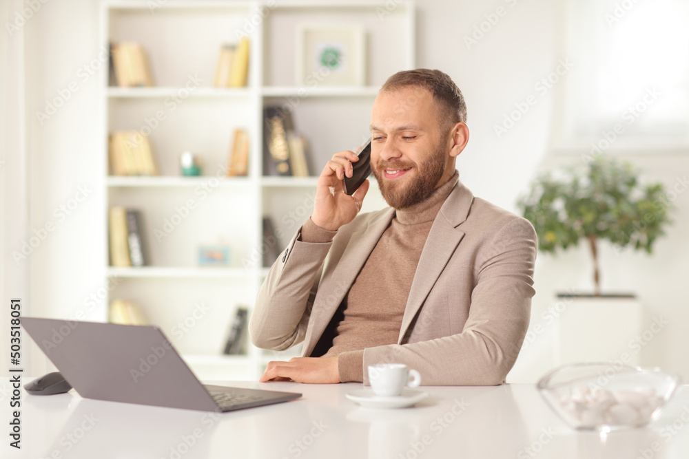 Happy young man sitting at home with a computer on a table and making a phone call Happy young man sitting at home with a computer on a table and making a phone call