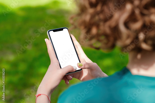 Young woman or teen girl holding smart phone mock up blank white screen in hand using mobile app at city park, outdoor. Order, read news, watch video, chatting or texting. Over shoulder closeup view.