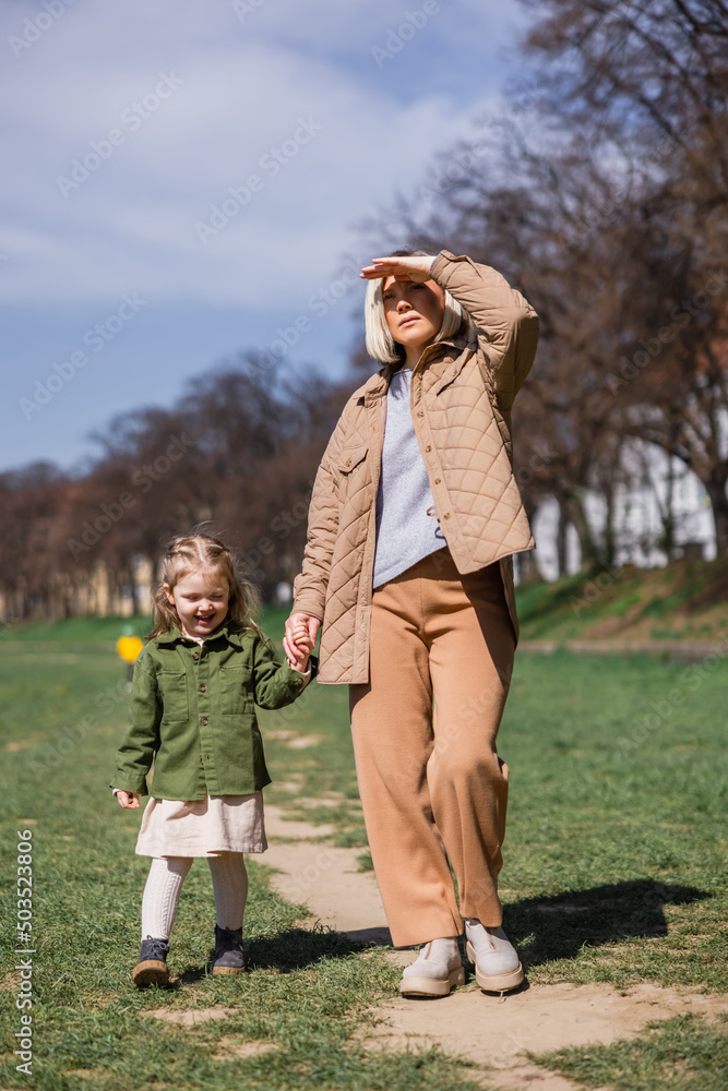 full length view of trendy woman looking away while walking with daughter in park. full length view of trendy woman looking away while walking with daughter in park.