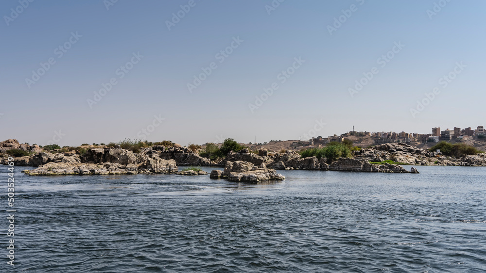 There are piles of picturesque boulders on the river bank. City houses are visible in the distance. Ripples on the blue water. Clear sky. Egypt. Nile There are piles of picturesque boulders on the river bank. City houses are visible in the distance. Ripples on the blue water. Clear sky. Egypt. Nile