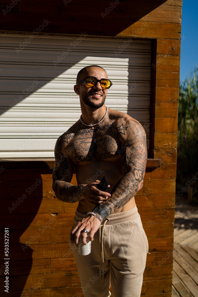 Chico joven rapado con tatuajes y musculado posando con ropa actual en la calle con mucho sol