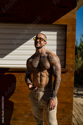 Chico joven rapado con tatuajes y musculado posando con ropa actual en la calle con mucho sol © MiguelAngelJunquera