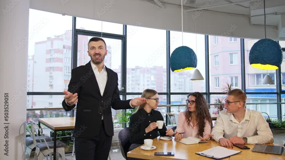 Businessman giving a talk to the camera and gesturing with his hands. Team of workmates sit next at the table having a conversation. Businessman giving a talk to the camera and gesturing with his hands. Team of workmates sit next at the table having a conversation.
