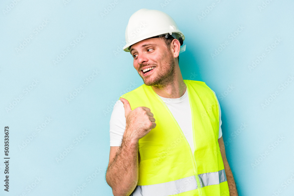 Young laborer caucasian man isolated on blue background points with thumb finger away, laughing and carefree.