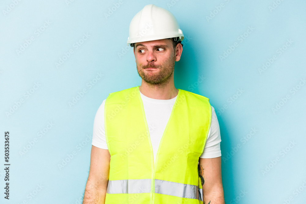 Young laborer caucasian man isolated on blue background confused, feels doubtful and unsure.