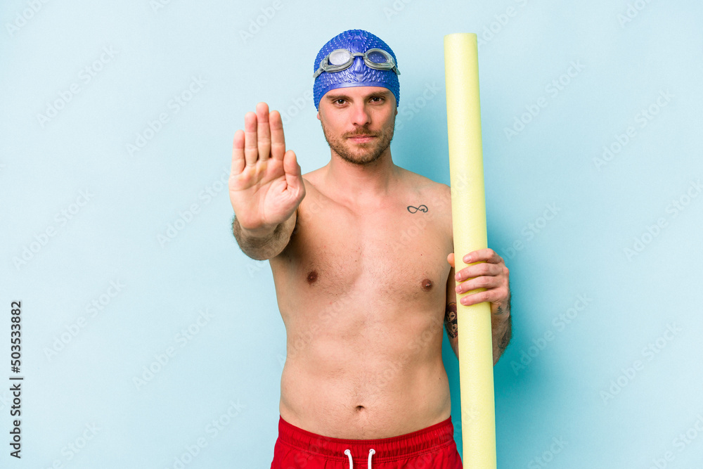 Young caucasian swimmer man holding foam stick isolated on blue background standing with outstretched hand showing stop sign, preventing you.