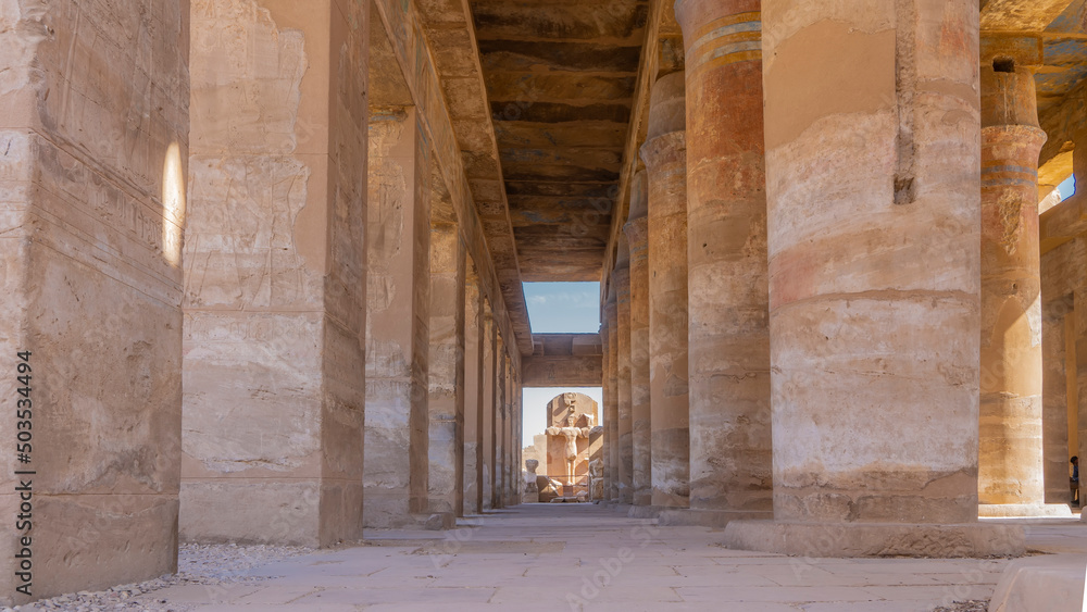 Smooth rows of columns in the Karnak temple of Luxor. Ahead, in the gap between the columns, a dilapidated statue is visible against the blue sky. Egypt