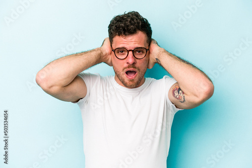 Young caucasian man isolated on blue background covering ears with hands trying not to hear too loud sound. © Asier