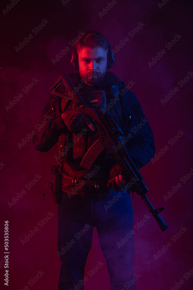 soldier in full gear with weapons. a man in headphones, body armor, with a backpack and a belt. red background. colored, blue-red light. smoke around the military. explosion, chemical attack