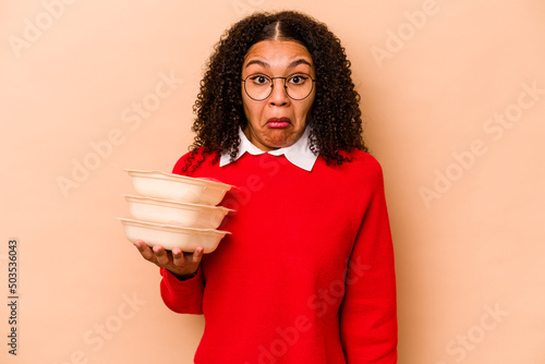 Young African American woman holding tupperware isolated on beige background shrugs shoulders and open eyes confused.