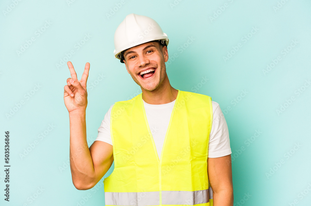 Young laborer caucasian man isolated on blue background joyful and carefree showing a peace symbol with fingers. Young laborer caucasian man isolated on blue background joyful and carefree showing a peace symbol with fingers.