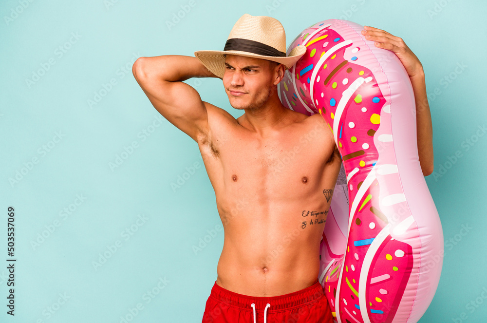 Young caucasian man holding an inflatable donut isolated on blue background touching back of head, thinking and making a choice.