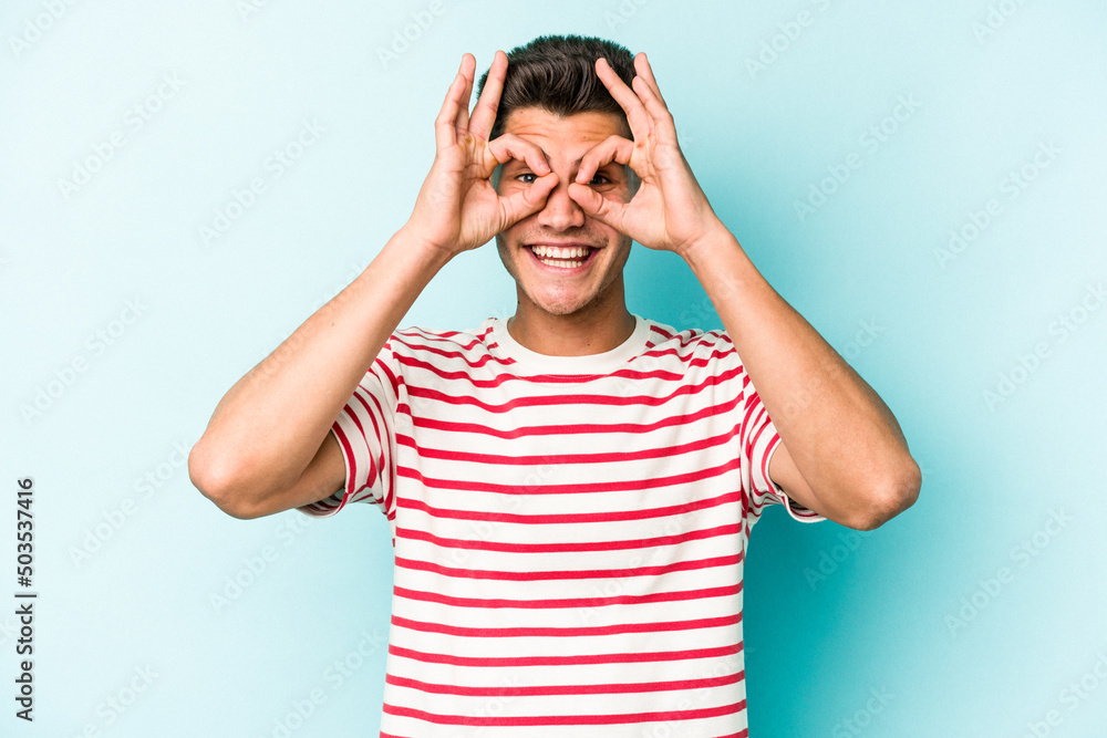 Young caucasian man isolated on blue background showing okay sign over eyes Young caucasian man isolated on blue background showing okay sign over eyes