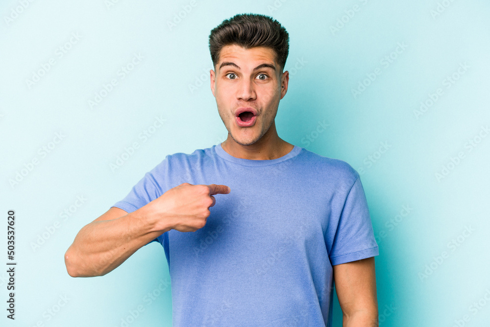 Young caucasian man isolated on blue background surprised pointing with finger, smiling broadly. Young caucasian man isolated on blue background surprised pointing with finger, smiling broadly.