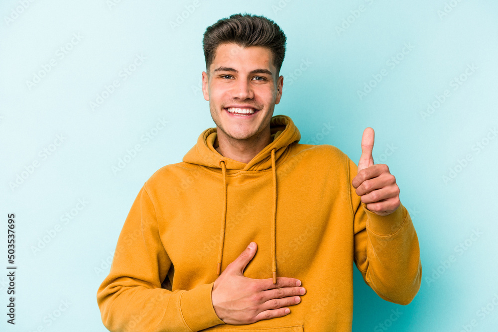 Fototapeta premium Young caucasian man isolated on blue background touches tummy, smiles gently, eating and satisfaction concept.