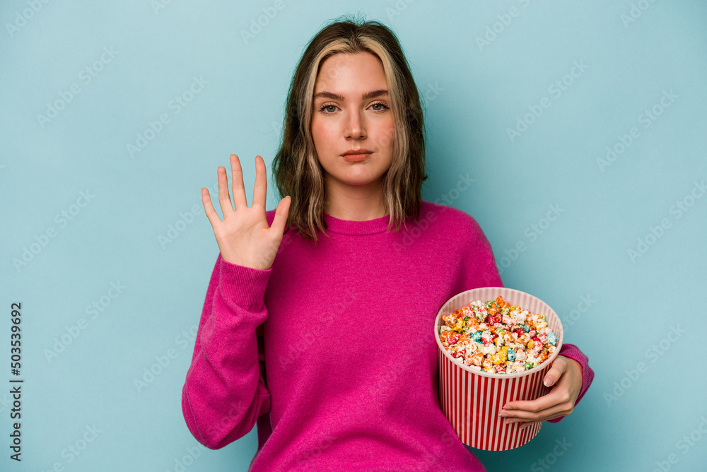Young caucasian woman holding popcorn isolated on blue background smiling cheerful showing number five with fingers.