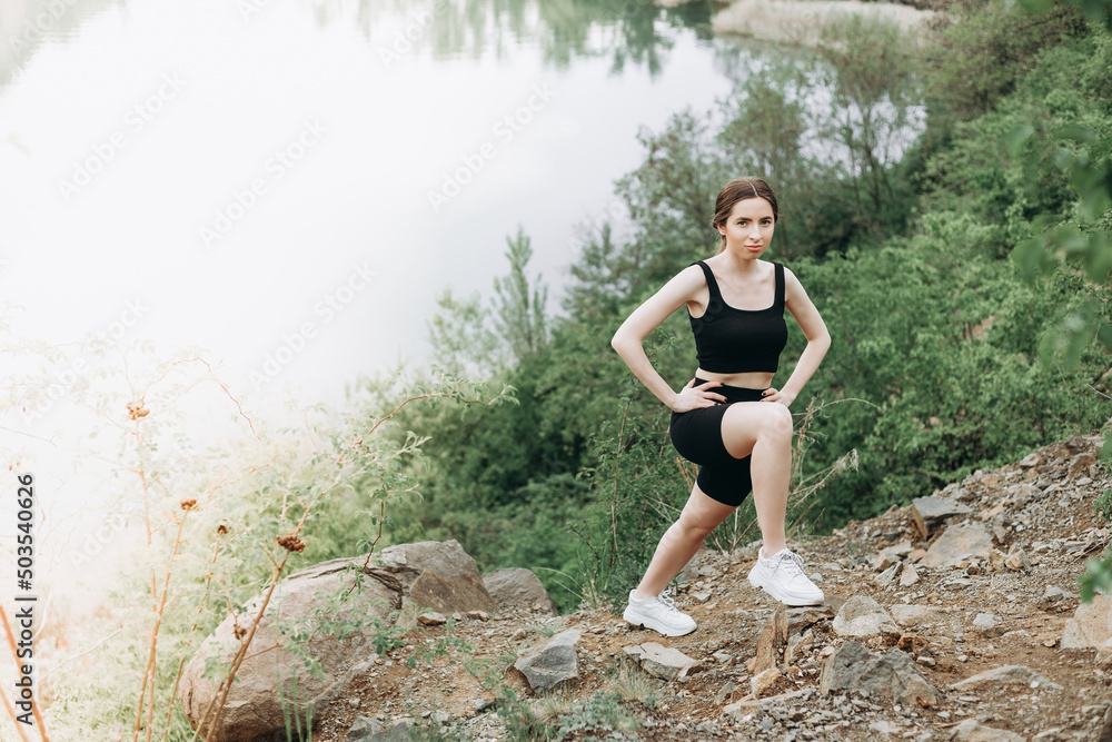 Young female runner stretching arms and legs before running at morning forest trail near lake