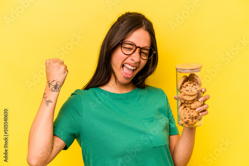 Canvas Young hispanic woman holding a cookies jar isolated on yellow background raising fist after a victory, winner concept