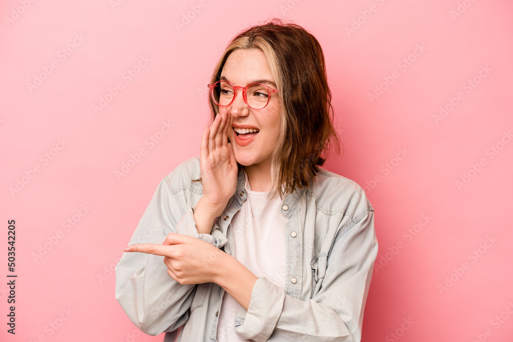 Young caucasian woman isolated on pink background saying a gossip, pointing to side reporting something.