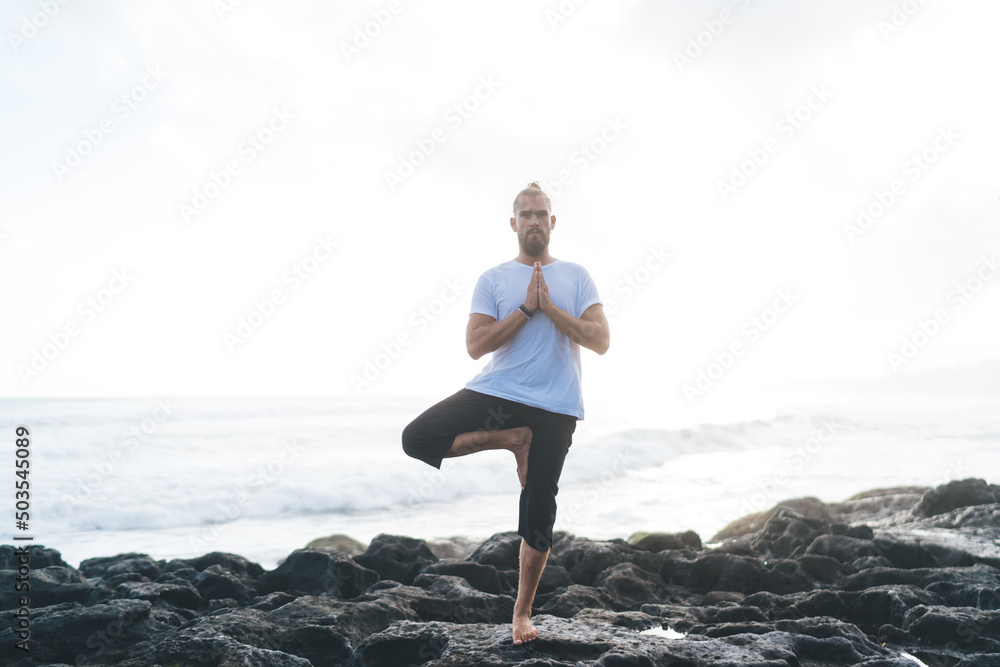 Caucasian man in casual sportswear doing asana poses for recreating during time for feeling mindfulness and appeasement, flexible male healing during holistic retreat training at coastline
