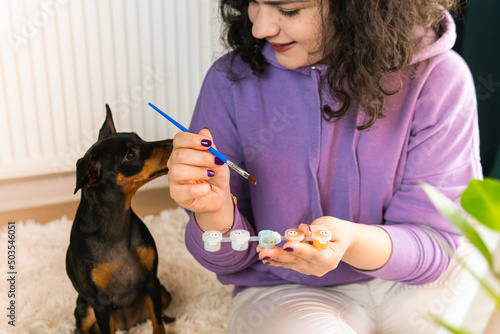 The adult woman painting by numbers indoors, her pet miniature pincher near. People with pets.