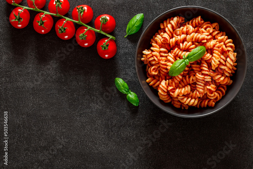 Fusilli, cooked pasta with tomato sauce, tomatoes cherry and basil leaf, on dark background, top view, space to copy text.