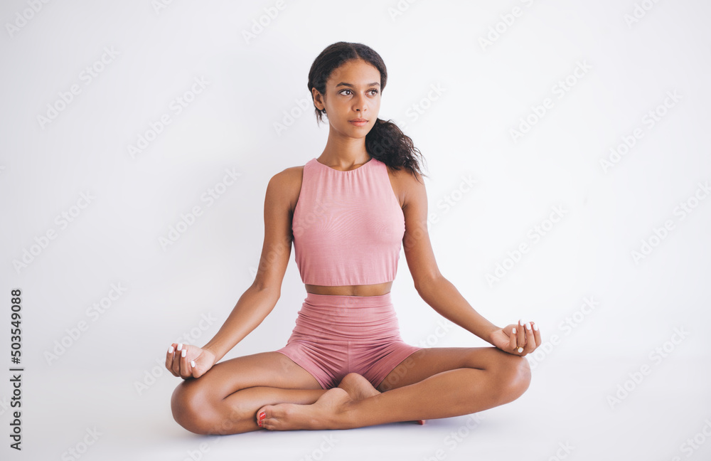 Harmony and meditation training during morning yoga in white gym studio, Dark skinned female in sportswear sitting in lotus pose during hatha practice for exercising body concentration in asana Harmony and meditation training during morning yoga in white gym studio, Dark skinned female in sportswear sitting in lotus pose during hatha practice for exercising body concentration in asana