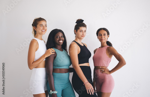 Group portrait of happy diverse fit girls of different race and body types in sportswear posing together, multiracial women in tracksuits smiling at camera against white background with copy space