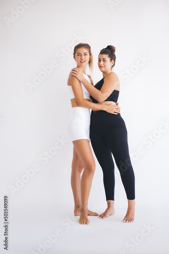 Full length portrait of Caucaisan female yoga partners in sportswear keeping healthy lifestyle, slim fit girls in tracksuit looking at camera during sportive photo session in white gym studio