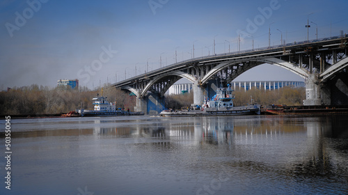 Two tugs pushing barges diverge under different spans of the bridge. River navigation with barges on a large river in the city.