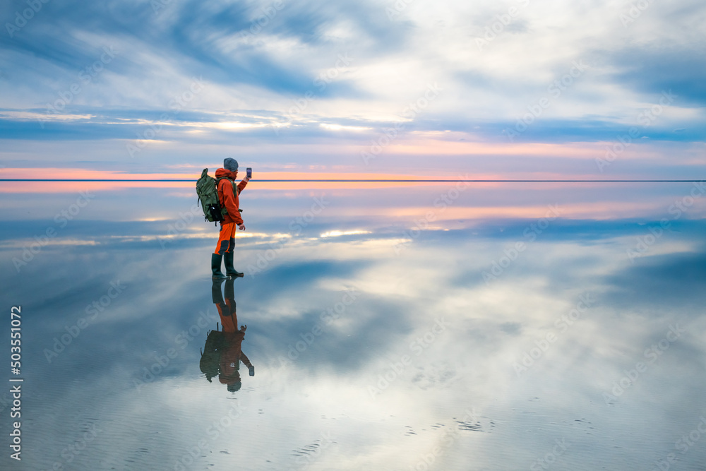 Man traveler with backpack taking photo with smartphone of the beautiful salt lake at sunset. Sky with clouds are reflected in the mirror water surface. Travel and adventure concept