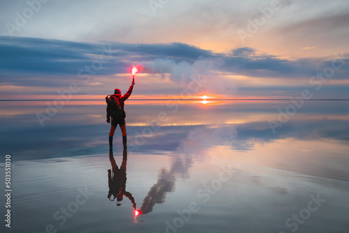 Man traveler stands on the salt lake at sunset and holds burning signal fire in his hand. Blue sky with clouds are reflected in the mirror water surface. Travel and adventure concept